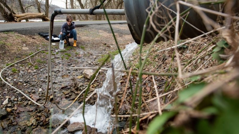 water coming from a pipe with a woman collecting samples in the background